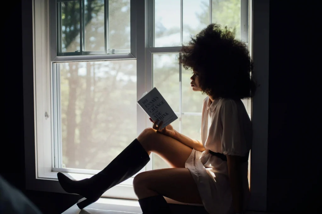 Imagem de uma mulher sentada em uma janela lendo um livro.