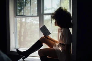 Imagem de uma mulher sentada em uma janela lendo um livro.